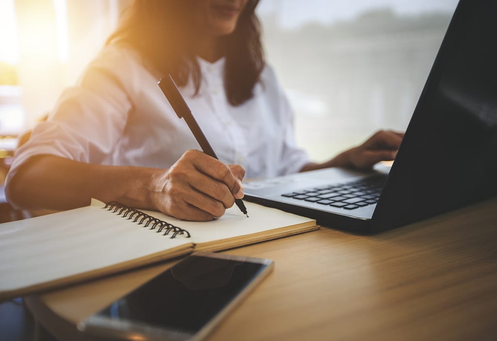 Woman focusing on her online business. Half of face is showing with hand on laptop, other hand holding pen, and writing on notebook with a phone in front.