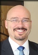 drgard | Walker Road Chiropractic Portrait of a smiling bald man with glasses in a dark suit and blue tie.