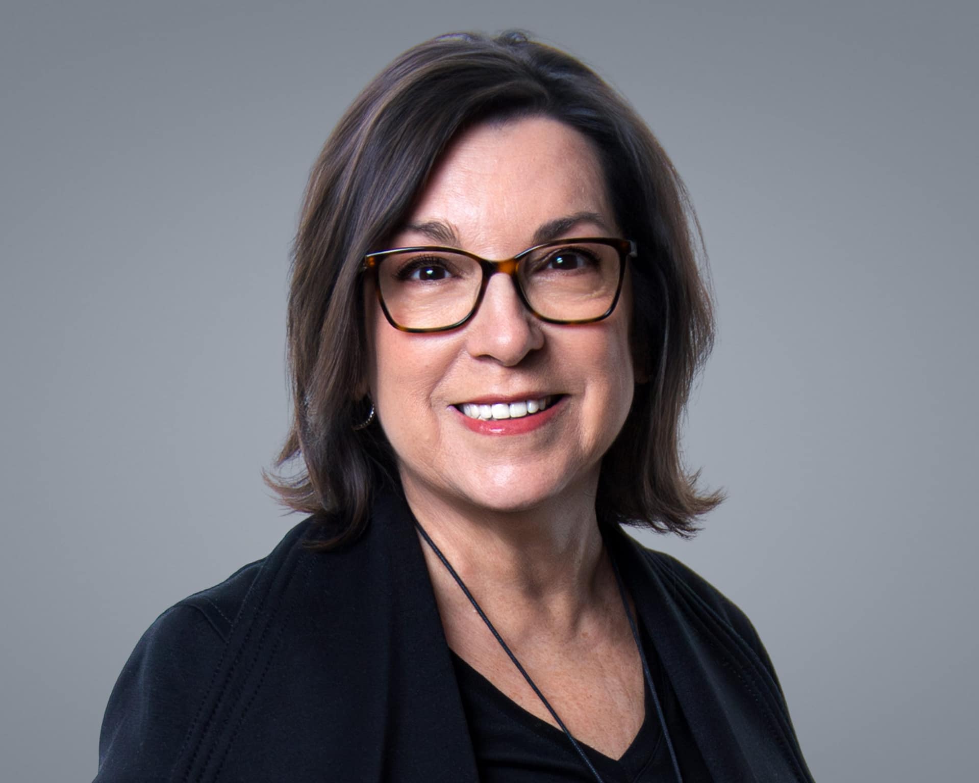 Ascione-Hilton P401p Holiday Planit Headshots-0219-Edit-2-4k | Holiday Planit Smiling woman with brown hair and tortoiseshell glasses, wearing a dark top against a gray background.