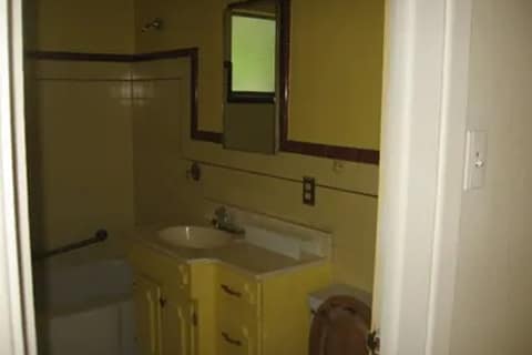 3a-411w | Advance Installations Tiny bathroom with a yellow vanity under a mirror, sink in front, and a tub/shower to the left. Window light in the back, wall switch nearby.