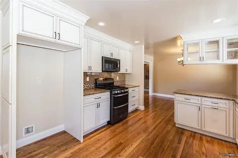 gallery1-a | Advance Installations Bright white kitchen with white cabinets, granite countertops, and a black freestanding stove on warm wood flooring.