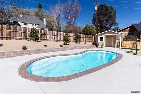 gallery10-a | Advance Installations Kidney-shaped in-ground pool in a sunny backyard with brick coping, concrete decking, a small shed, and a wooden fence behind.