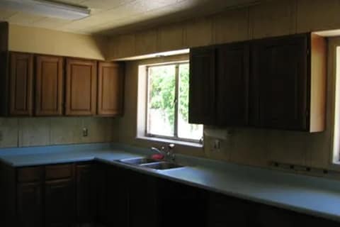 gallery2-b | Advance Installations Dim kitchen with dark wood cabinets, pale countertops, and a double sink under a window; greenery visible outside.