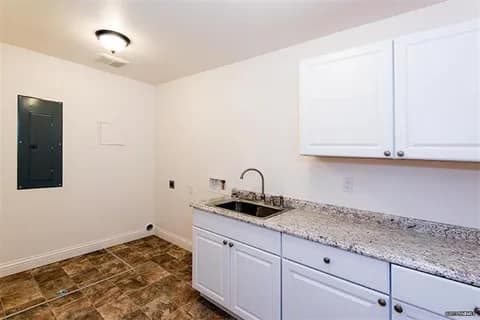gallery7-a | Advance Installations A small white kitchen or utility area with granite counter, sink, white cabinets, and brown tiled floor, plus an electrical panel on the left wall and bright ceiling light