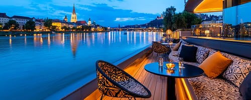 Luxurious riverboat deck at dusk with cushioned seating, a round table with candles, and city lights reflecting on calm water behind.