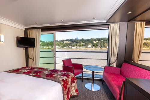 Hotel room with a bed in foreground, red seating, and a round blue table; large windows overlook a river and tree-covered hills outside.