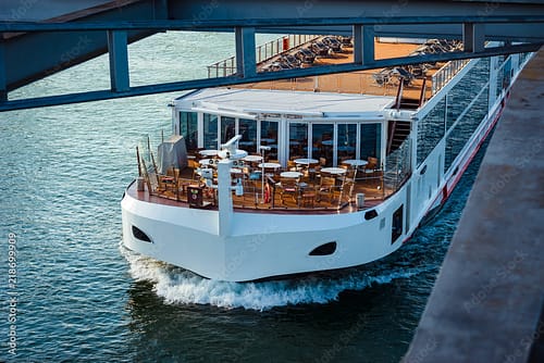 White cruise boat cruising under a bridge, with outdoor dining on the upper deck and lounge chairs visible on the sun deck.