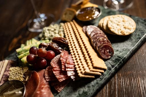 Somersett Food Commercial Final 17 Assorted charcuterie on a slate board: sliced salami and cured meats, grapes, cucumber sticks, crackers, bread rounds, and a small bowl of dip with a few nuts nearby