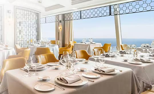 Vista-overview-dining-concept-left-1920x580 | Holiday Planit Elegant seaside restaurant with white-tablecloth tables set with plates, glassware, and yellow chairs, overlooking the ocean