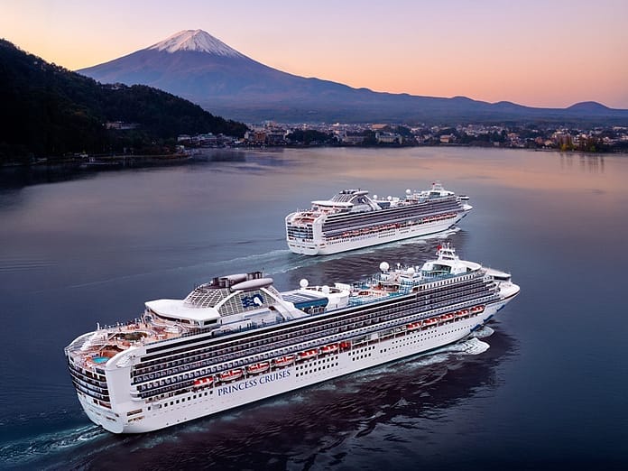 default | Holiday Planit Two large Princess Cruises ships sail on a calm bay at sunset with a mountain and town in the distance.
