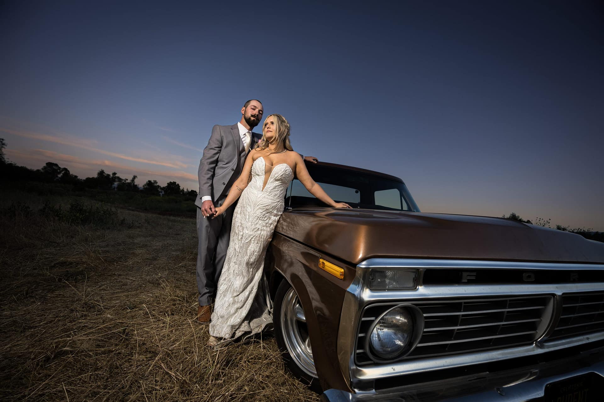 Bride in a white lace gown and groom in a gray suit lean on a vintage brown pickup against a dusk sky in a dry field, wedding photo.