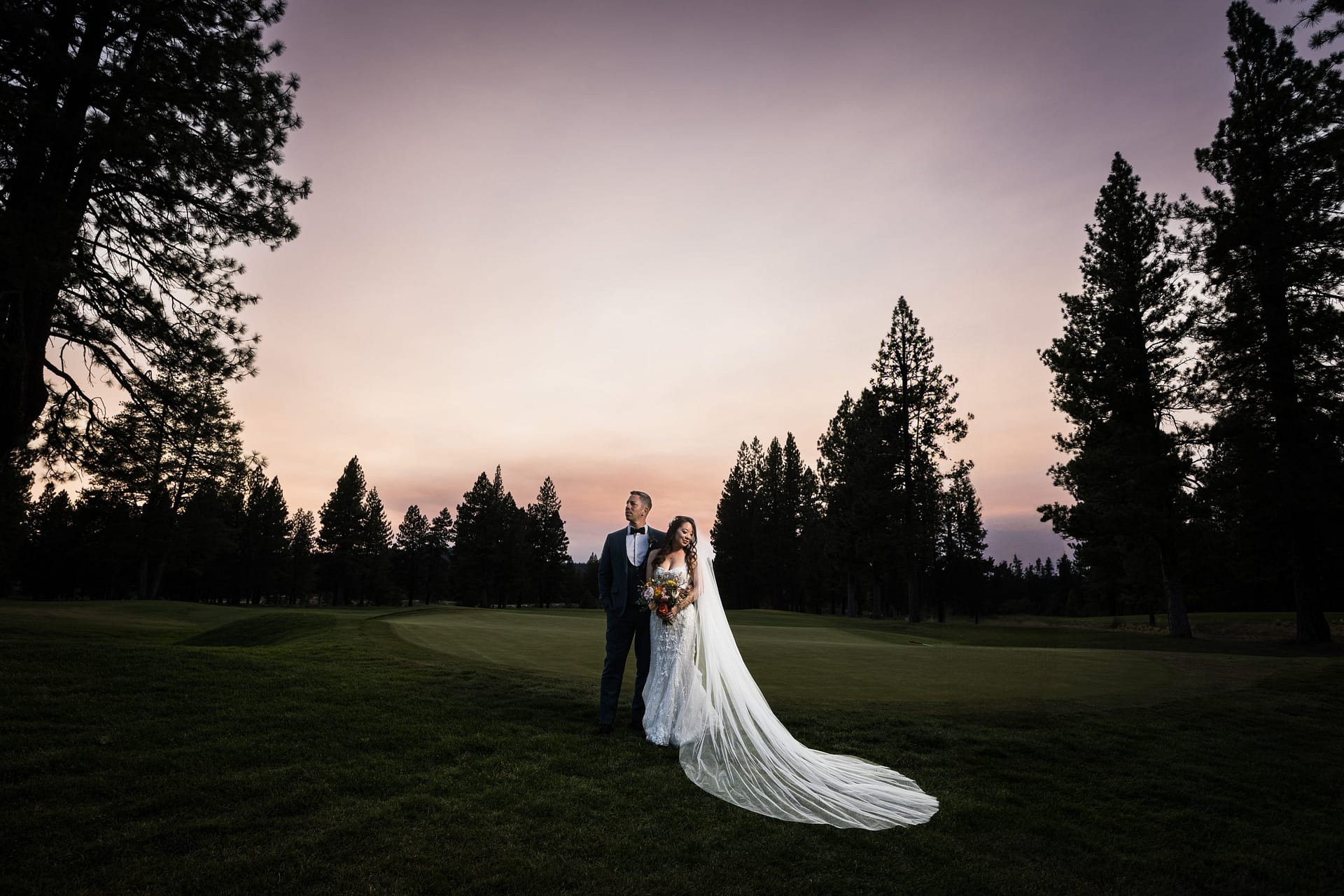 Bride in a white lace gown with a long veil holds a bouquet beside a groom in a dark suit on a grassy field at sunset, with tall pine trees framing the scene.
