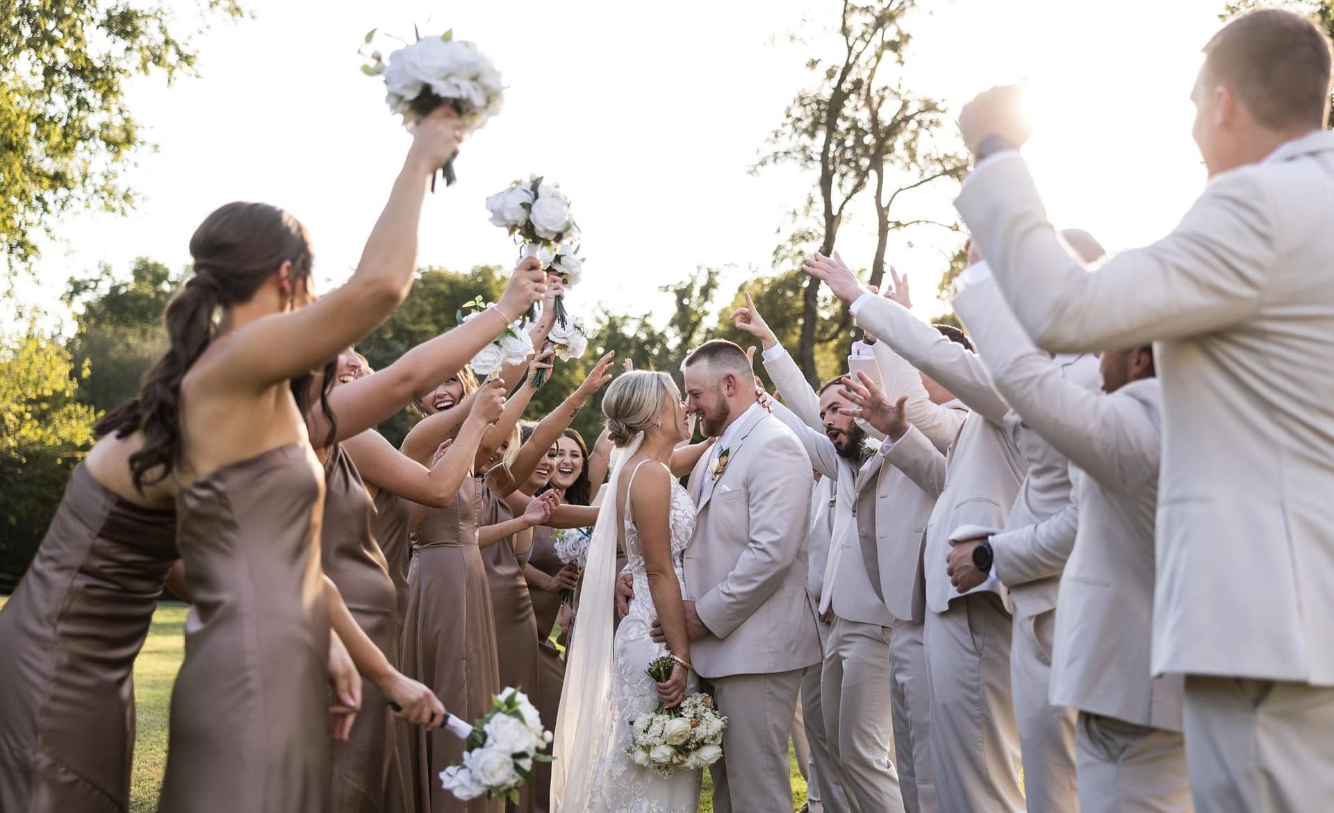 Bride and groom kiss as bridal party in taupe dresses and gray suits cheerfully raise bouquets outdoors.
